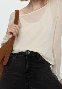 Personne portant un haut beige transparent à manches longues et un jean noir taille haute, tenant la sangle d'un sac à bandoulière en cuir marron avec la main gauche.