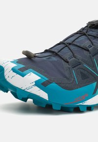 Chaussure de trail running avec une tige bleu marine foncé, des accents turquoise, une surface texturée, une semelle agressive et un marquage Contagrip sur le côté.