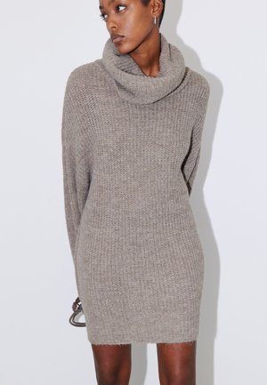 Vrouw draagt een losse, lange mouwen, taupekleurige gebreide sweaterjurk met een grote col, staand tegen een effen witte achtergrond.