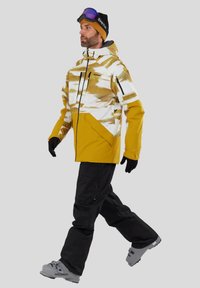 Gul og hvid mønstret skijakke med sorte detaljer, kombineret med sorte bukser og grå skistøvler, designet til vintersport.