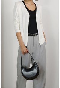 Borsa in pelle nera a forma di mezzaluna con una tracolla curva, abbinata a un cardigan bianco, una maglia nera e pantaloni eleganti grigi.