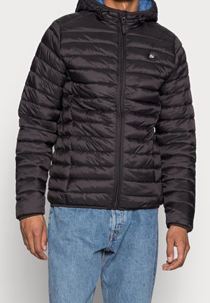 Mand iført en sort quiltet hættetrøjejakke med lynlås foran og lyseblå jeans, stående mod en neutral baggrund.