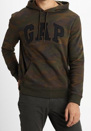 Mann trägt grünen und braunen camouflagegemusterten GAP-Hoodie mit schwarzen Kordeln und dunklen Hosen, steht mit einer Hand in der Tasche.