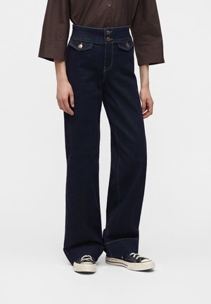 Person iført mørkeblå vidde jeans med høj talje, to forlommer med knapper foran og sorte lave sneakers med hvide såler.