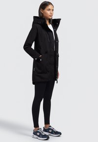 Parka noir longue avec capuche, fermeture zip, deux grandes poches avant et une texture résistante à l'eau, associée à des leggings noirs et des chaussures de sport.