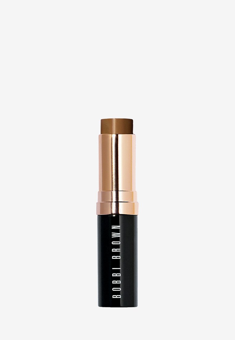 Foundationstick mit goldfarbenem Metallkopf und schwarzem zylindrischem Sockel. Das Etikett lautet "BOBBI BROWN." Braune Cremeprodukte sind oben sichtbar.