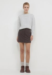 Frau mit kurzen Haaren, die einen hellgrauen, strukturierten Pullover, einen braunen, gestreiften Minirock, beigefarbene Socken und braune Stiefeletten trägt und an einer weißen Wand steht.