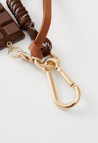 Sangle en nylon marron avec des accents blancs, arborant un charm en forme de barre chocolatée. Fermeture en métal doré pour l'attache. Finitions lisses et texturées.