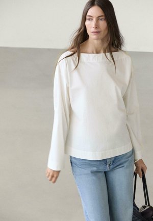 Kobieta z długimi brązowymi włosami, ubrana w biały długi rękaw i jasnoniebieskie jeansy, trzymająca czarną plecioną torbę w lewej ręce.