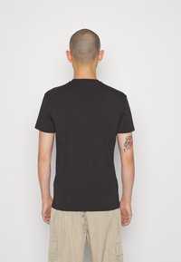 T-shirt in cotone nero con maniche corte e scollatura rotonda, vista dal retro. Presenta un tatuaggio sul braccio destro.