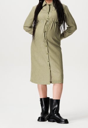 Person mit langen Zöpfen, die ein grün-weiß gestreiftes, geknöpftes Kleid und schwarze Lederstiefel bis zur Mitte der Wade trägt, steht mit den Händen hinter dem Rücken.