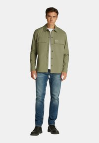 Camicia verde oliva con bottoni, dotata di due tasche sul petto, indossata sopra una t-shirt bianca, abbinata a jeans blu e stivali neri.