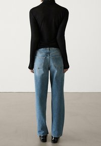 Vrouw in een zwart langmouwen shirt en lichtblauwe rechte jeans, staan op blote voeten op een grijze vloer tegen een effen witte muur.