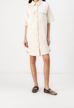 Femme portant une robe chemise crème à manches courtes et des ballerines imprimé léopard avec des brides noires sur fond blanc.