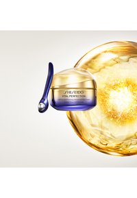 Shiseido Vital Perfection ögonkräm i en burk med ett gyllene lock och blå bas. I sällskap av en blå applicator med en sfärisk spets. Gyllene orb bakgrund.