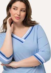 Blusa azul clara de cuello en V con mangas de tres cuartos, que presenta acentos a rayas en navy y blanco en los puños y el escote. Textura de tela suave.