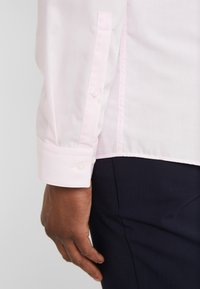 Camicia a maniche lunghe rosa chiaro con polsini a bottone, caratterizzata da una texture liscia e una vestibilità su misura. È abbinata a pantaloni blu scuro.