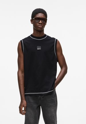 Homme portant un débardeur noir avec des coutures blanches, un jean foncé et des lunettes de soleil larges et foncées, debout avec une main dans la poche.