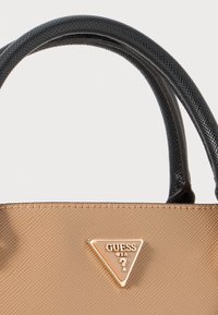 Primo piano di una borsa tan con manici neri texture e logo triangolare dorato Guess USA sul davanti.