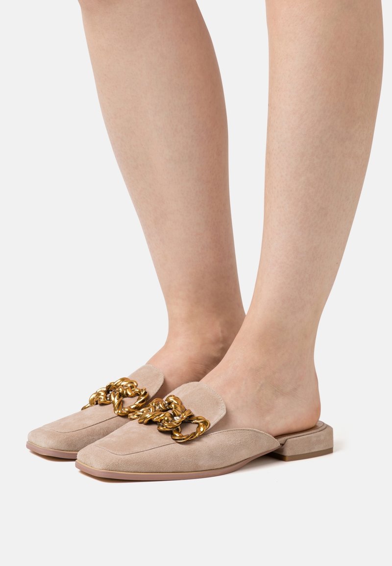 Mules en daim beige avec un talon bas et un accent décoratif en chaîne dorée sur la partie supérieure. Texture lisse avec un design à bout arrondi.