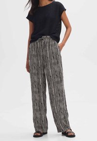 Donna che indossa pantaloni larghi a righe verticali bianchi e neri, una maglietta nera a maniche corte e sandali neri da infilare, in piedi con una mano in tasca.