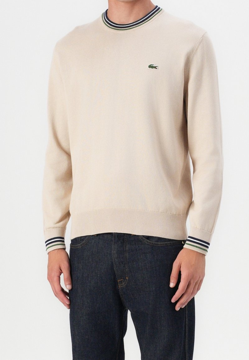 Pull beige à manches longues avec col et poignets rayés, arborant un petit logo crocodile vert sur la poitrine, porté avec un jean bleu foncé.