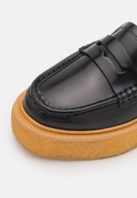 Mocassin en cuir noir avec semelle en caoutchouc épaisse texturée couleur noisette, couture détaillée, et fente pour pièce sur le dessus avant.
