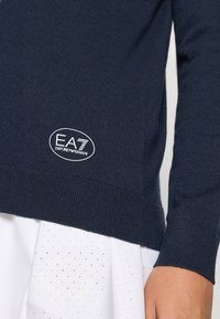 Námornícky pletený sveter s rebrovanými rukávmi a spodným okrajom, s bielou nášivkou loga EA7 Emporio Armani na dolnej ľavej strane.