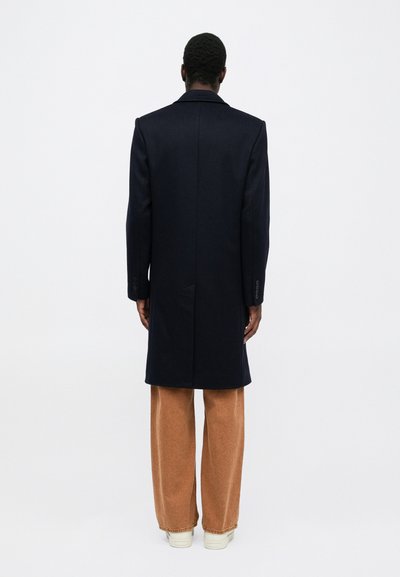 Marineblauer Wollmantel mit geradem Schnitt, einfachem Schlitz und geknöpften Manschetten, kombiniert mit hellbraunen weiten Hosen und weißen Turnschuhen.