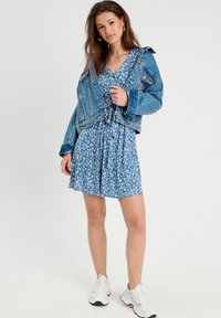 Chaqueta de denim sobre un vestido floral azul. El vestido tiene mangas cortas y una falda acampanada. Los accesorios incluyen pendientes de aro y zapatillas deportivas blancas.
