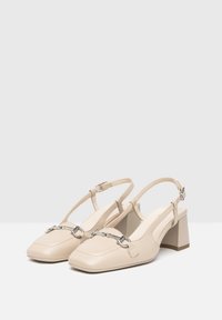 Tacchi slingback beige con punta quadrata, caratterizzati da un accentuato con catena nella parte anteriore e un tacco spesso. Realizzati in pelle liscia con finitura opaca.