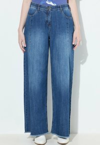 Mujer vestida con jeans de pierna ancha de talle alto en azul con dobladillo sin terminar y zapatos blancos de punta, de pie frente a un fondo blanco.