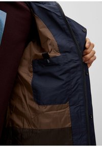 Laivastonsininen takki, jossa on ruskea vuori, sisällä painonappitasku ja vetoketjullinen suljenta, osittain auki, jotta sisäpuoli näkyy.