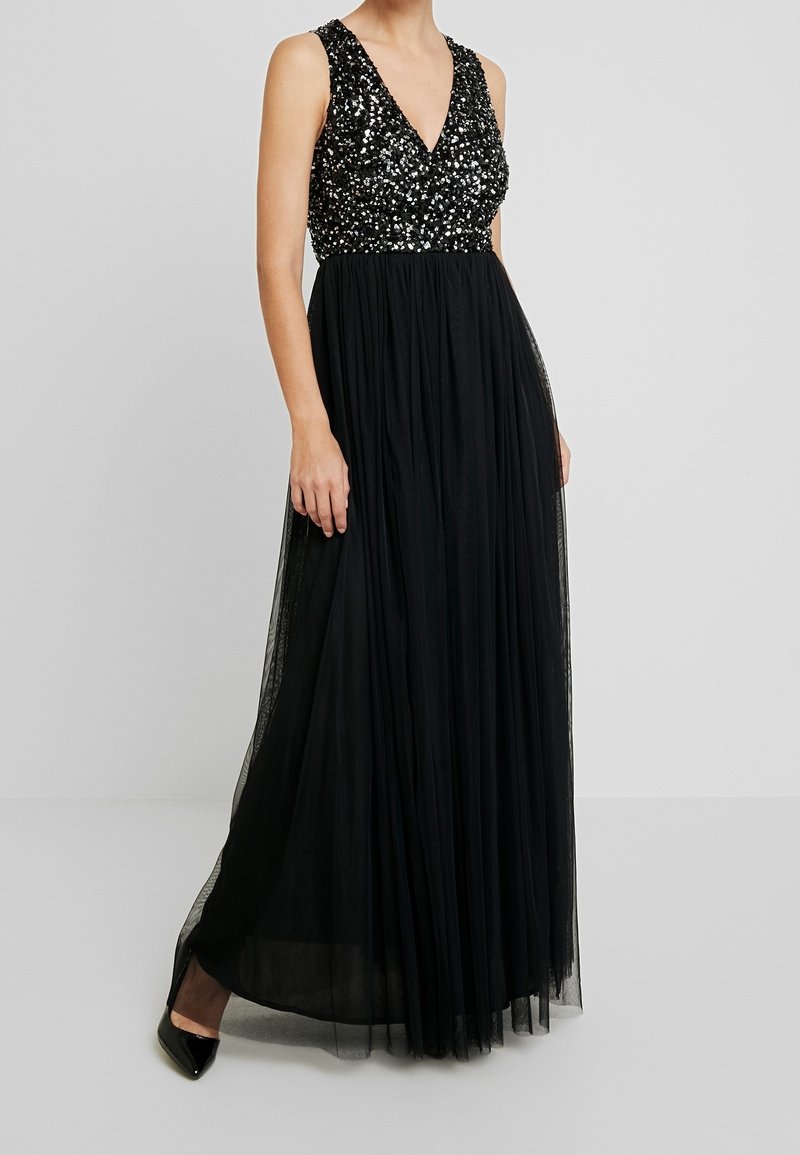Femme portant une robe noire sans manches avec un corsage à sequins et une jupe fluide transparente, associée à des escarpins noirs à bout pointu.