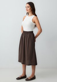 Top bianco senza maniche abbinato a una gonna marrone scuro di lunghezza al ginocchio con una cintura elastica. Scarpe nere piatte con un design ritagliato.