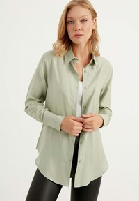 Camicia a maniche lunghe verde chiaro con frontale a bottoni, colletto e orlo curvo. Tessuto morbido e design leggero, indossata sopra un top bianco.