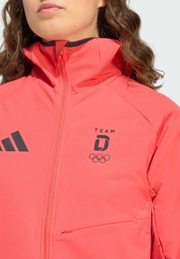 Koraalrode sportjas met hoge kraag, ritssluiting, twee zakken, zwarte logo's van Adidas en Olympische ringen, gladde stoftextuur.
