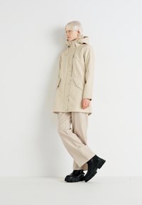Parka beige imperméable avec une capuche, fermeture à pression à l'avant et deux poches latérales, associée à un pantalon beige clair et des bottes noires chunky.