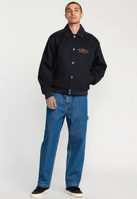 Black bomber jacket with orange logo text, paired with loose-fitting blue jeans and black slip-on shoes against a neutral background.