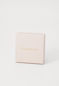 Coccinelle-laatikko pehmeässä vaaleanpunaisessa pahvissa, jossa on mattapinta ja kultakoristeltu logo etupuolella. Neliön muotoinen, yksinkertainen muotoilu.