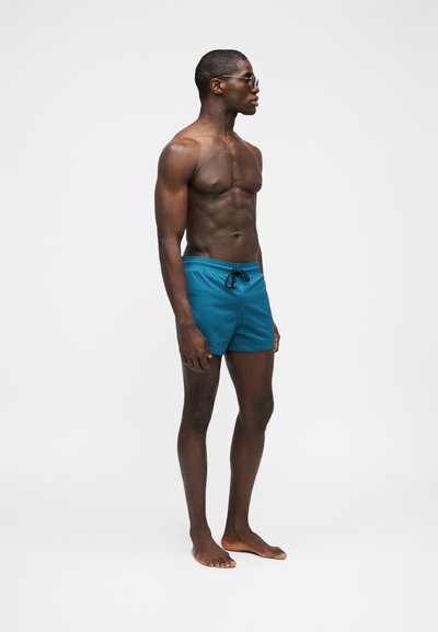 Μυώδης άντρας φορώντας πράσινο-μπλε μαγιό και γυαλιά ηλίου, όρθιος ξυπόλητος, κοιτάζοντας προς τα δεξιά με φόντο ένα απλό λευκό τοίχο.