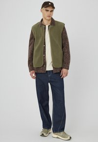 Gilet en polaire vert par-dessus une chemise à motif marron, un T-shirt crème, un pantalon en denim bleu et des chaussures de sport beige. Il porte une casquette à l'envers.