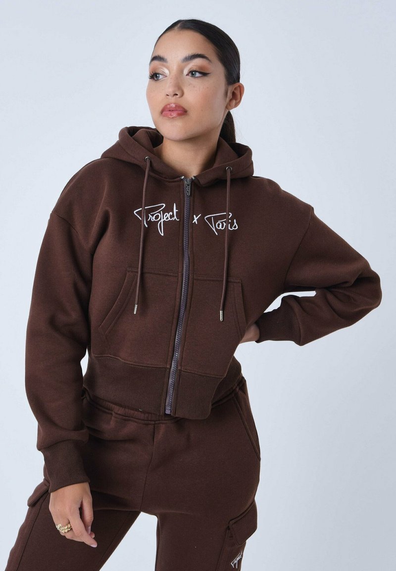 Haut hoodie court marron avec fermeture éclair, présentant des poches avant et le texte "Project x Paris" en broderie blanche. Texture du tissu lisse.