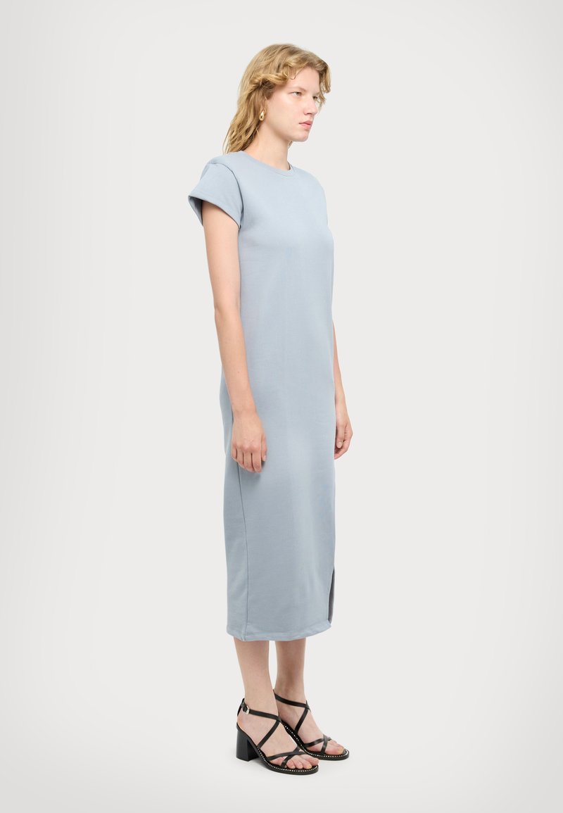 Vestido azul claro, hasta la rodilla, con mangas cortas enrolladas, un escote redondo y una abertura lateral. Llevado con tacones negros con tiras.