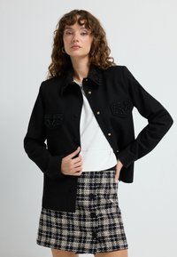 Black jacket with beaded collar and pockets, featuring gold buttons. Worn over a white top and paired with a black and white plaid skirt.