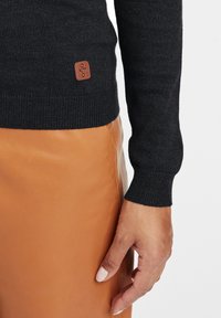 Zwarte gebreide trui met geribde manchetten, voorzien van een klein bruin leren label. Gecombineerd met een gladde oranje leren rok.