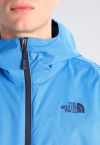 Ljusblå jacka med dragkedja framtill, som har en huva. Inkluderar en svart logotyp på bröstet. Tillverkad av ett strukturerat, ventilerande tyg.