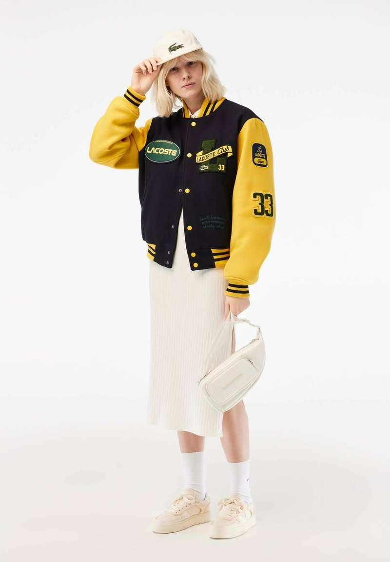 Veste varsity Lacoste noire et jaune avec des écussons, présentant des poignets côtelés, une jupe côtelée blanche et des baskets crème. Sac à main blanc.