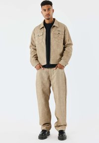 Chaqueta de sarga en color canela y pantalones a juego, que presentan dos bolsillos en el pecho, cierre con cremallera y un tejido texturizado, combinados con zapatos negros.
