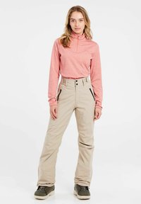 Lichtroze shirt met lange mouwen en een halve ritssluiting, gecombineerd met beige sneeuwbroeken met zijritsen en zwarte accenten, gedragen met groene laarzen.